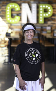 A smiling woman in a visor and Chicken N Pickle t-shirt stands indoors. Large illuminated letters CNP are visible on the wall behind her.