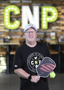 A smiling man in a CNP shirt and hat holds a pickleball paddle and ball. Large illuminated CNP letters are displayed in the background.