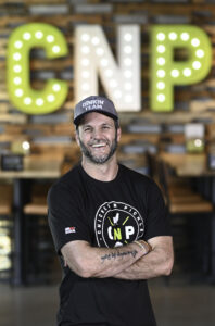 A smiling man in a Dinkin Team cap and Chicken N Pickle shirt stands with arms crossed in front of large lit letters CNP on a brick wall.