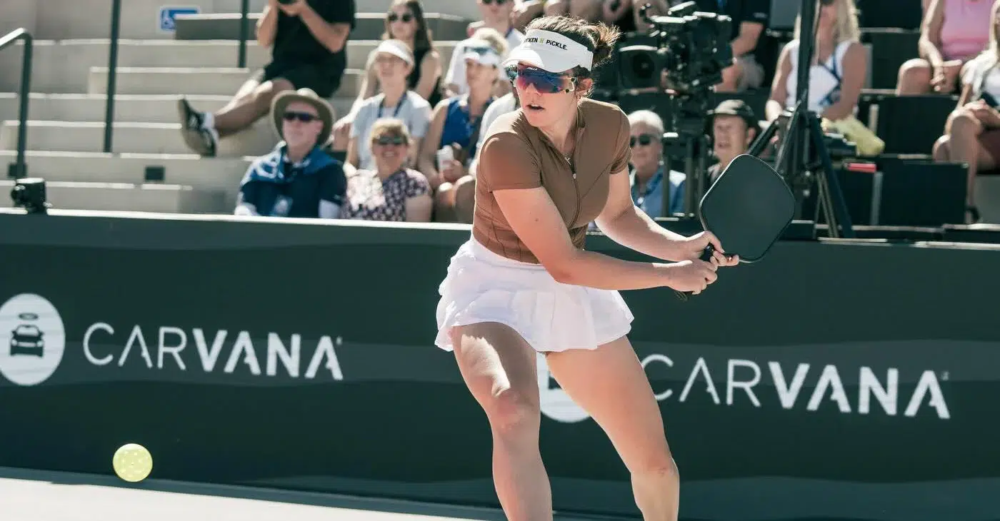 A woman wearing a visor and sunglasses prepares to hit a pickleball on an outdoor court, with a crowd watching in the background.