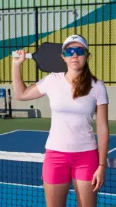 A woman in a pink outfit and visor stands on a pickleball court, holding a paddle over her shoulder. She wears blue sports sunglasses and looks confidently at the camera. The court and net are visible behind her.