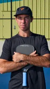 A man in a black shirt and hat stands on a blue court, arms crossed, holding a PIKKL pickleball paddle, with a chain-link fence and yellow wall in the background.