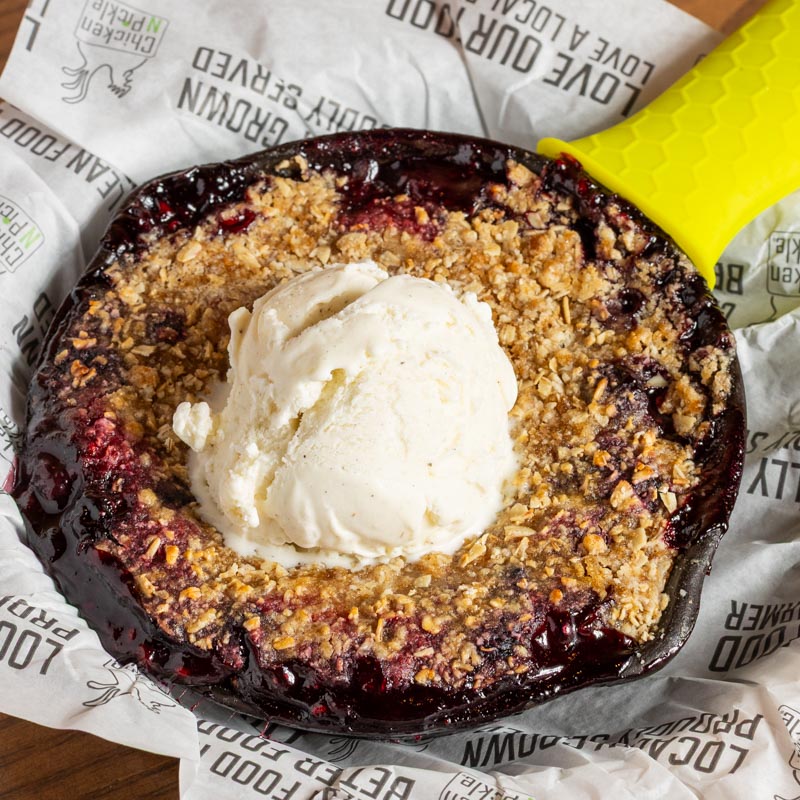 A small skillet filled with a berry crumble dessert, topped with a scoop of vanilla ice cream, sits on branded parchment paper.
