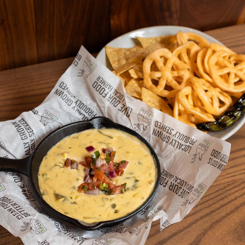 A small skillet of queso dip topped with chopped tomatoes, onions, and herbs sits beside a plate of tortilla chips and wagon wheel chips on a wooden table.