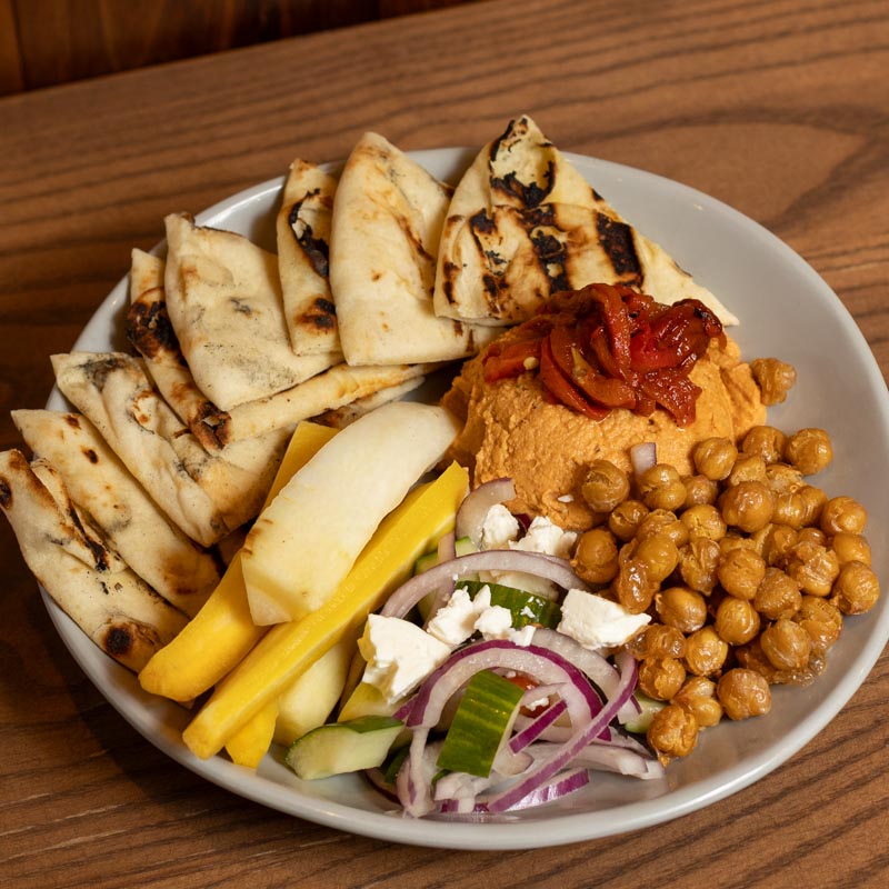 A plate with grilled pita slices, hummus topped with red peppers, roasted chickpeas, sliced yellow and white vegetables, and a cucumber, onion, and feta salad, all arranged neatly on a wooden table.