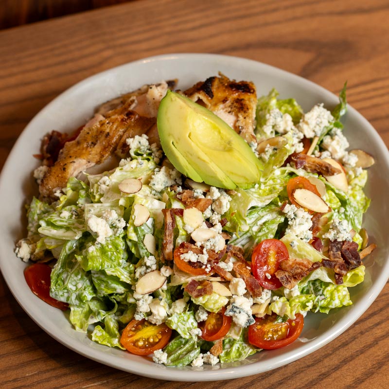 A bowl of salad with grilled chicken, avocado slices, cherry tomatoes, blue cheese crumbles, bacon bits, sliced almonds, and chopped romaine lettuce on a wooden table.