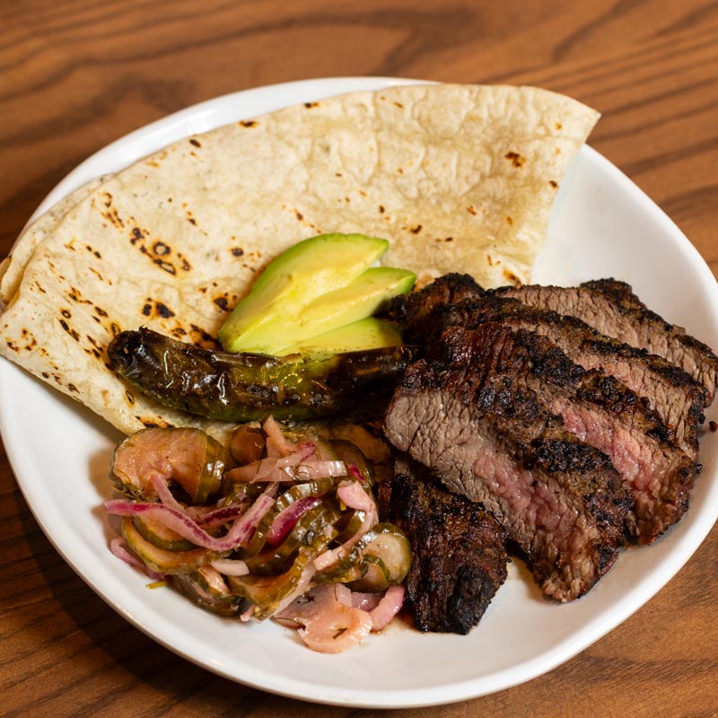 A white plate with grilled steak slices, a flour tortilla, avocado slices, grilled pepper, and a pickled onion and cucumber mix on a wooden table.