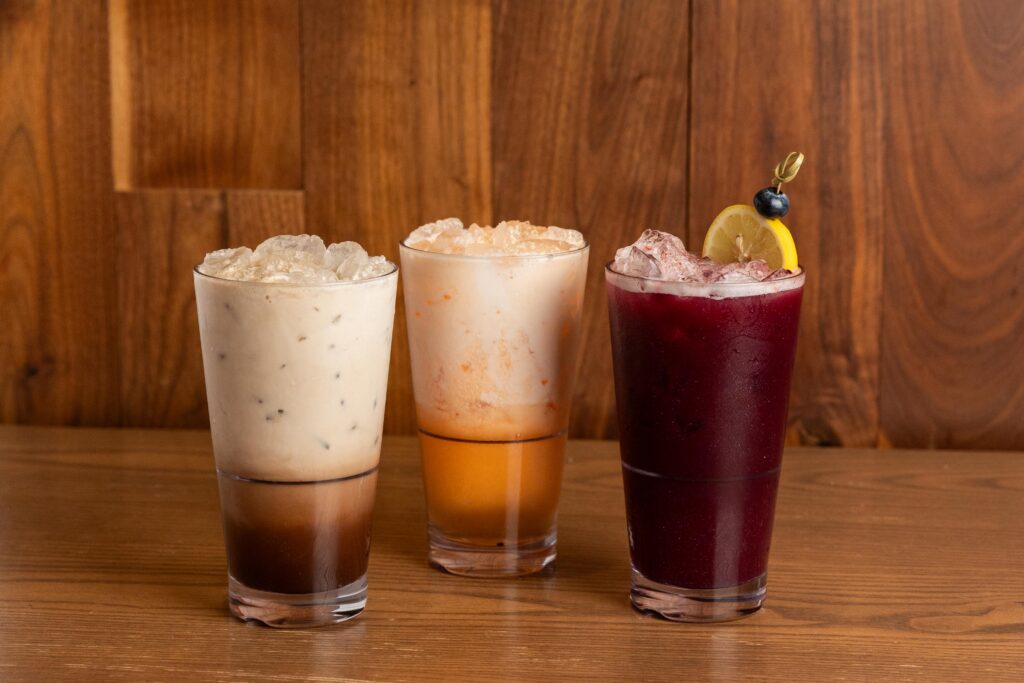 Three iced drinks in clear glasses sit on a wooden table. The drinks are brown with cream, orange and cream, and dark purple with lemon and blueberry garnish. A wood panel wall is in the background.