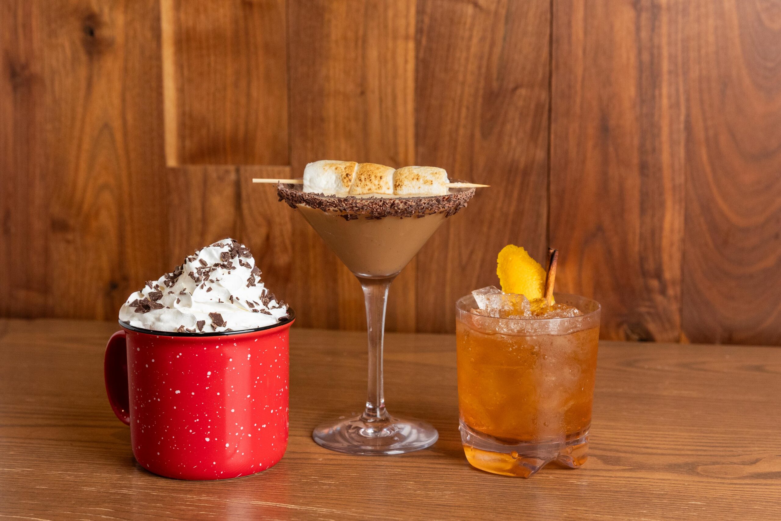 Three drinks on a wooden table: a red mug with whipped cream and chocolate shavings, a martini glass with a smores garnish, and a cocktail with ice, an orange slice, and a cherry.