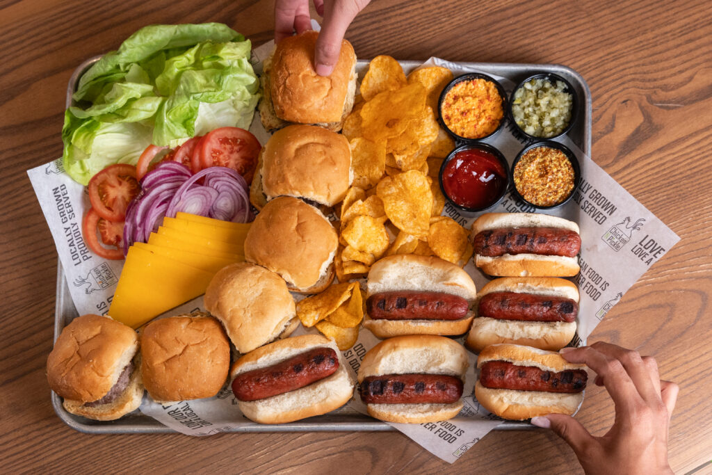 A tray with sliders, hot dogs, potato chips, sliced cheese, lettuce, tomato, onion, and condiments. Two hands are reaching for the food on a wooden table.