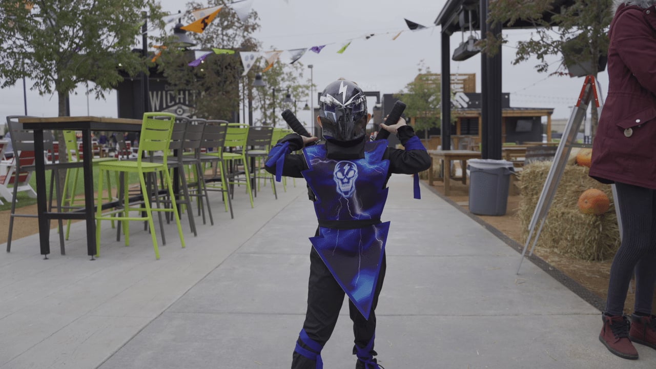 A child in a blue ninja costume poses with toy swords outdoors near green chairs, hay bales, and festive decorations.
