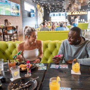 Two people sit at a table with food, drinks, and bingo cards, smiling and holding party poppers in a lively, modern restaurant with bright yellow seating and a BE AMAZING sign in the background.