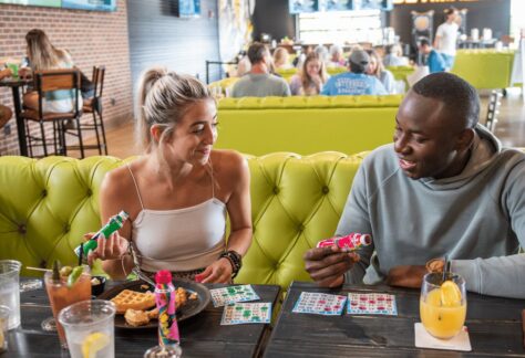 Two people sit at a table with food, drinks, and bingo cards, smiling and holding party poppers in a lively, modern restaurant with bright yellow seating and a BE AMAZING sign in the background.