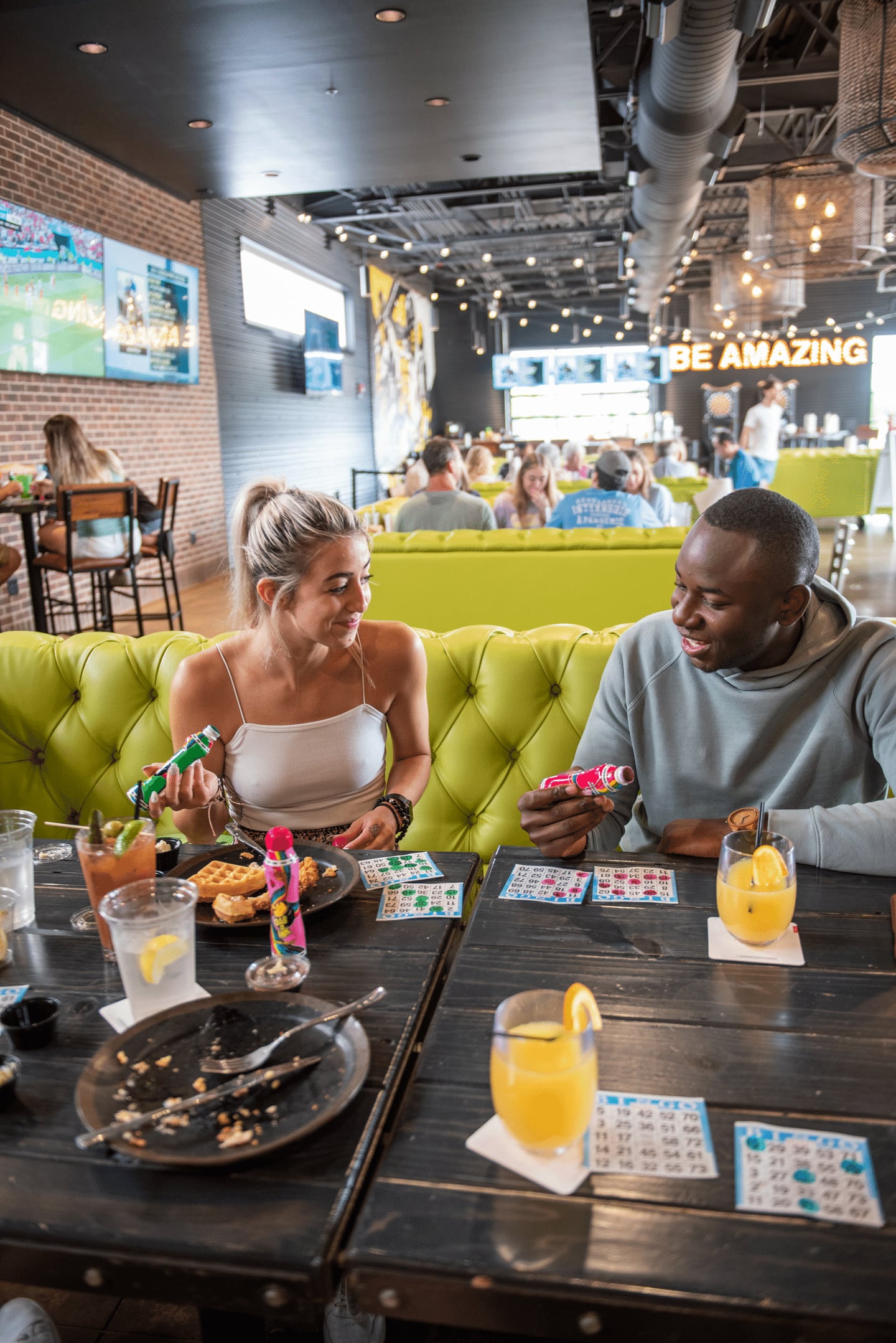 Two people sit at a table with food, drinks, and bingo cards, smiling and holding party poppers in a lively, modern restaurant with bright yellow seating and a BE AMAZING sign in the background.
