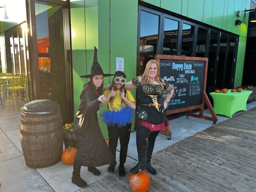 Three people in costumes—a witch, a minion, and a pirate—pose outside a green building with pumpkins and a chalkboard sign, enjoying a festive, daytime event.