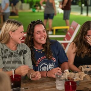 Three young women sit at a picnic table outdoors, laughing together. Food, drinks, and napkins are on the table. Other people are blurred in the background, suggesting a social gathering.