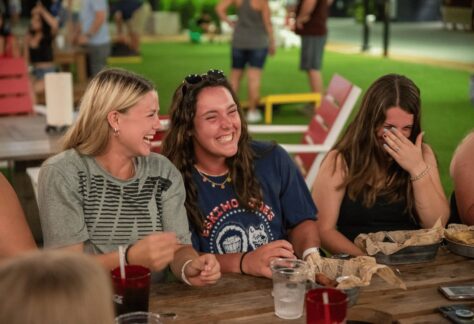 Three young women sit at a picnic table outdoors, laughing together. Food, drinks, and napkins are on the table. Other people are blurred in the background, suggesting a social gathering.