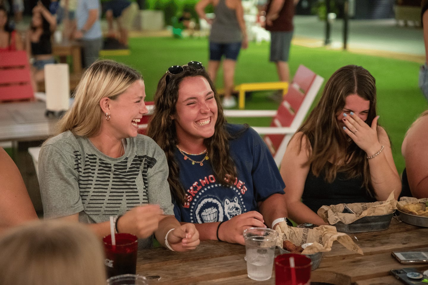 Three young women sit at a picnic table outdoors, laughing together. Food, drinks, and napkins are on the table. Other people are blurred in the background, suggesting a social gathering.