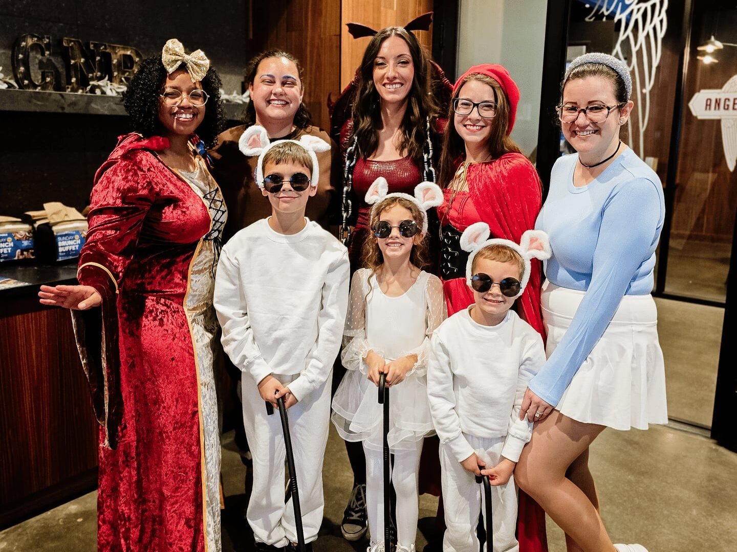A group of adults and children in creative costumes pose together indoors, smiling at the camera. The children wear matching white mouse outfits and sunglasses. The adults wear themed costumes, including fairy tale and character attire.