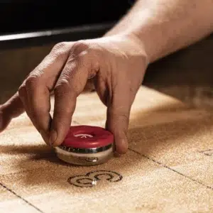 A hand prepares to slide a red shuffleboard puck across a wooden shuffleboard table, aiming for the scoring zones marked with numbers.