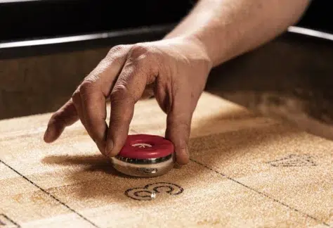 A hand prepares to slide a red shuffleboard puck across a wooden shuffleboard table, aiming for the scoring zones marked with numbers.