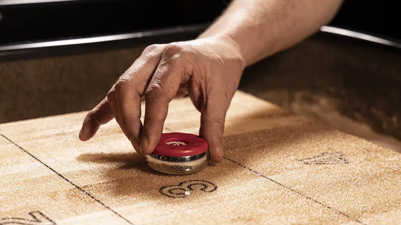 A hand prepares to slide a red shuffleboard puck across a wooden shuffleboard table, aiming for the scoring zones marked with numbers.