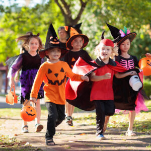 Six children in Halloween costumes, including witches, a pumpkin, a vampire, and a skeleton, run outdoors on a sunny day carrying orange pumpkin candy buckets.
