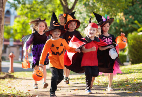 Six children in Halloween costumes, including witches, a pumpkin, a vampire, and a skeleton, run outdoors on a sunny day carrying orange pumpkin candy buckets.
