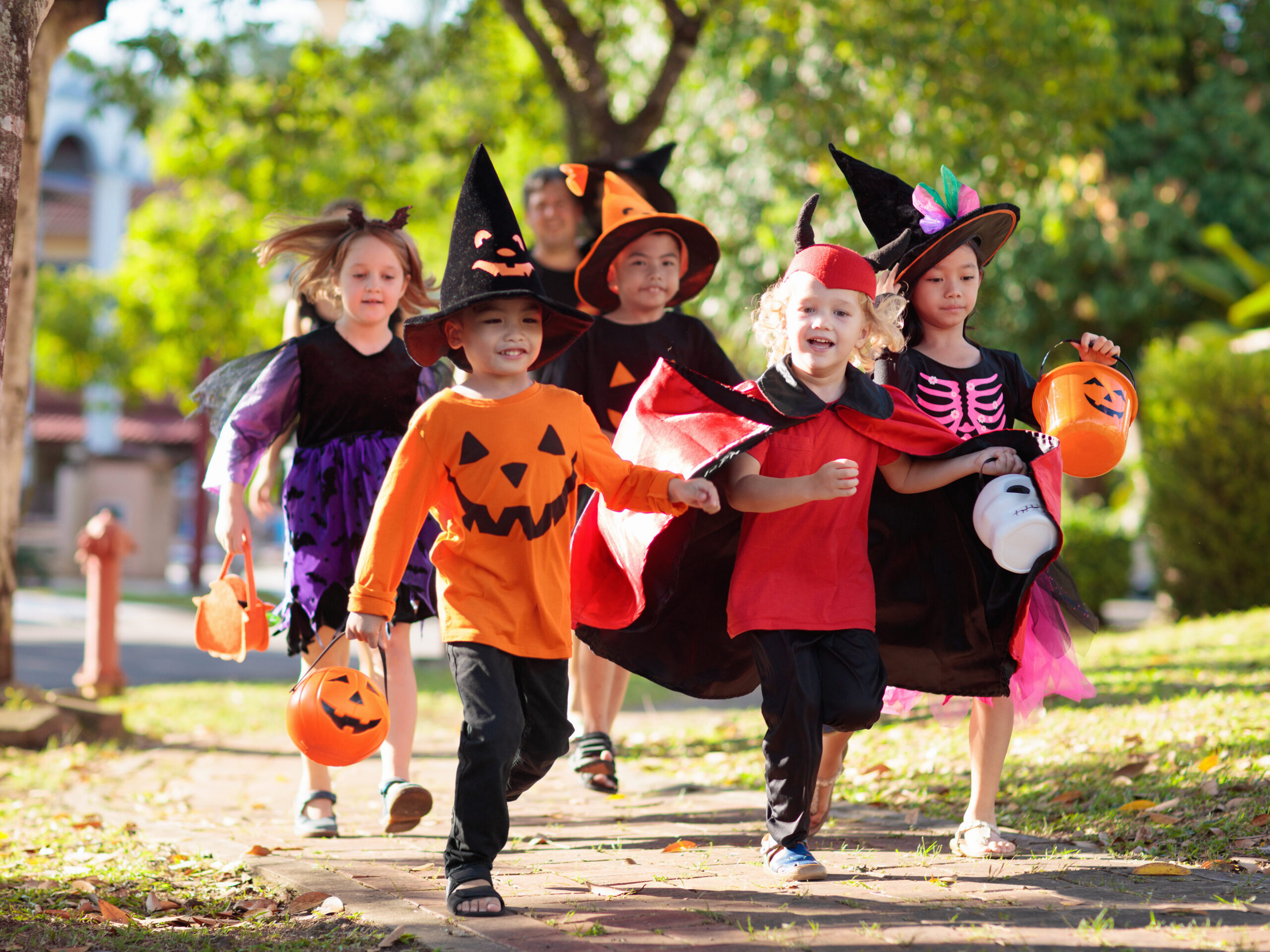 Six children in Halloween costumes, including witches, a pumpkin, a vampire, and a skeleton, run outdoors on a sunny day carrying orange pumpkin candy buckets.