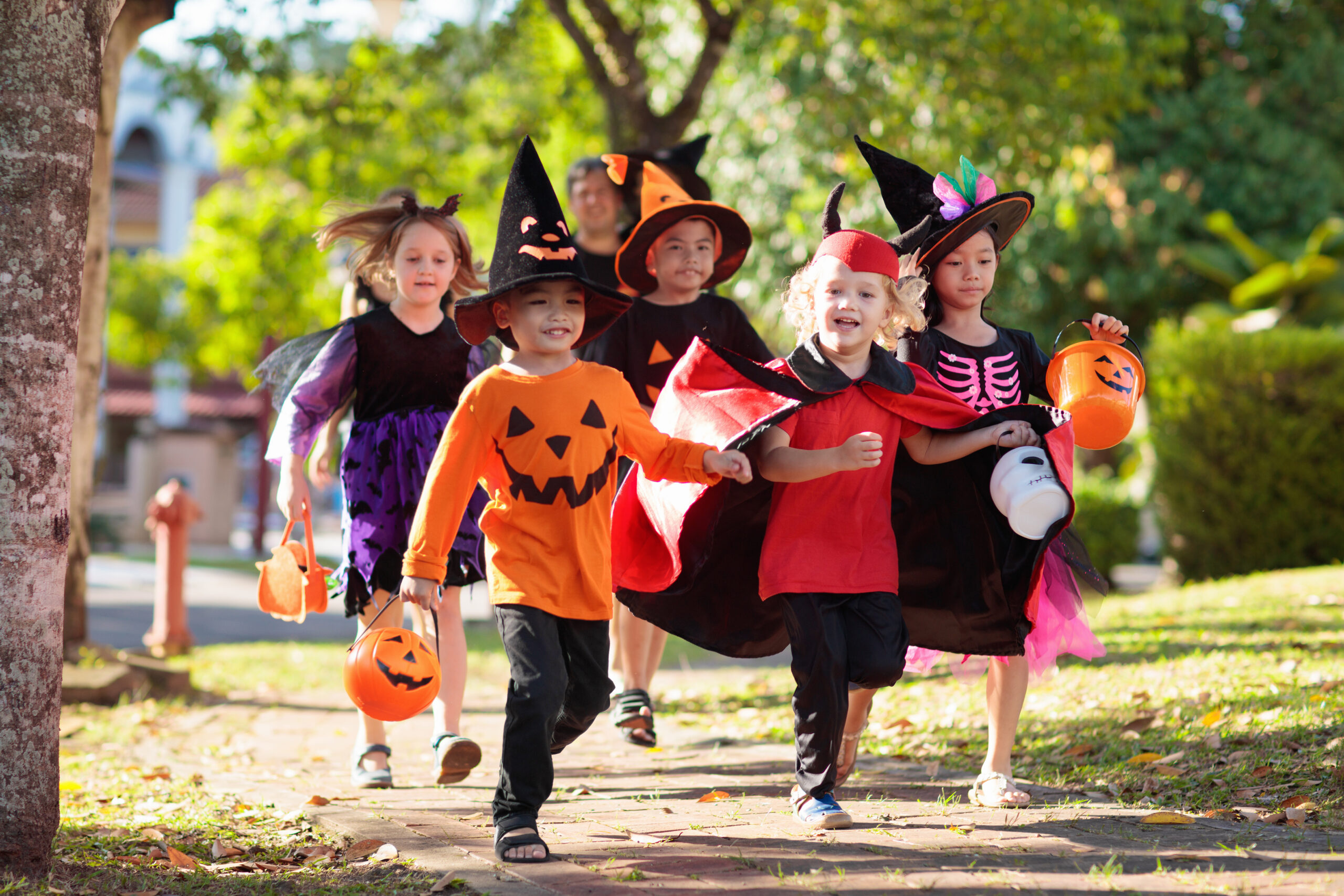 Six children in Halloween costumes, including witches, a pumpkin, a vampire, and a skeleton, run outdoors on a sunny day carrying orange pumpkin candy buckets.