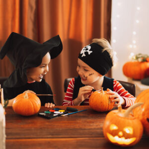 Two children in Halloween costumes paint small pumpkins at a table, surrounded by jack-o-lanterns and other pumpkins. They smile at each other, enjoying the festive activity in a cozy, decorated room.