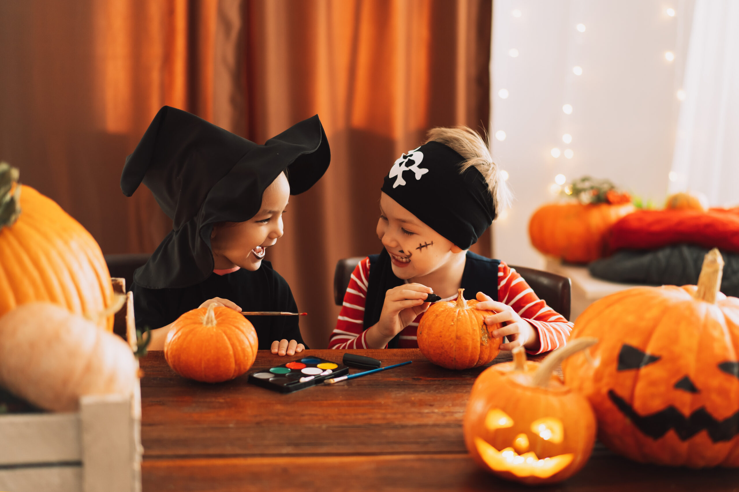 Two children in Halloween costumes paint small pumpkins at a table, surrounded by jack-o-lanterns and other pumpkins. They smile at each other, enjoying the festive activity in a cozy, decorated room.