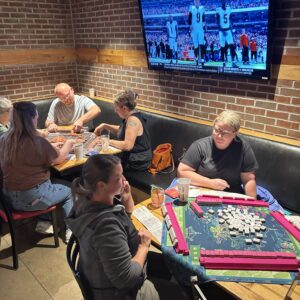 People play games at two tables in a restaurant with brick walls; one group is playing mahjong, and another is playing a different game. A TV above them shows a football game. Drinks and bags are on the tables and benches.