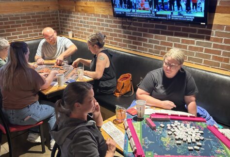People play games at two tables in a restaurant with brick walls; one group is playing mahjong, and another is playing a different game. A TV above them shows a football game. Drinks and bags are on the tables and benches.