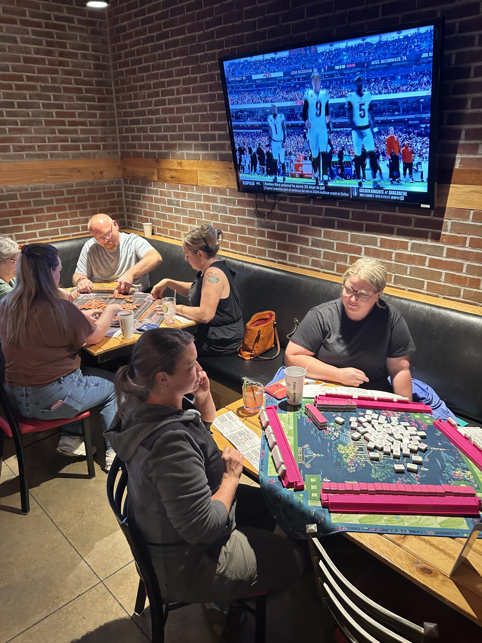 People play games at two tables in a restaurant with brick walls; one group is playing mahjong, and another is playing a different game. A TV above them shows a football game. Drinks and bags are on the tables and benches.