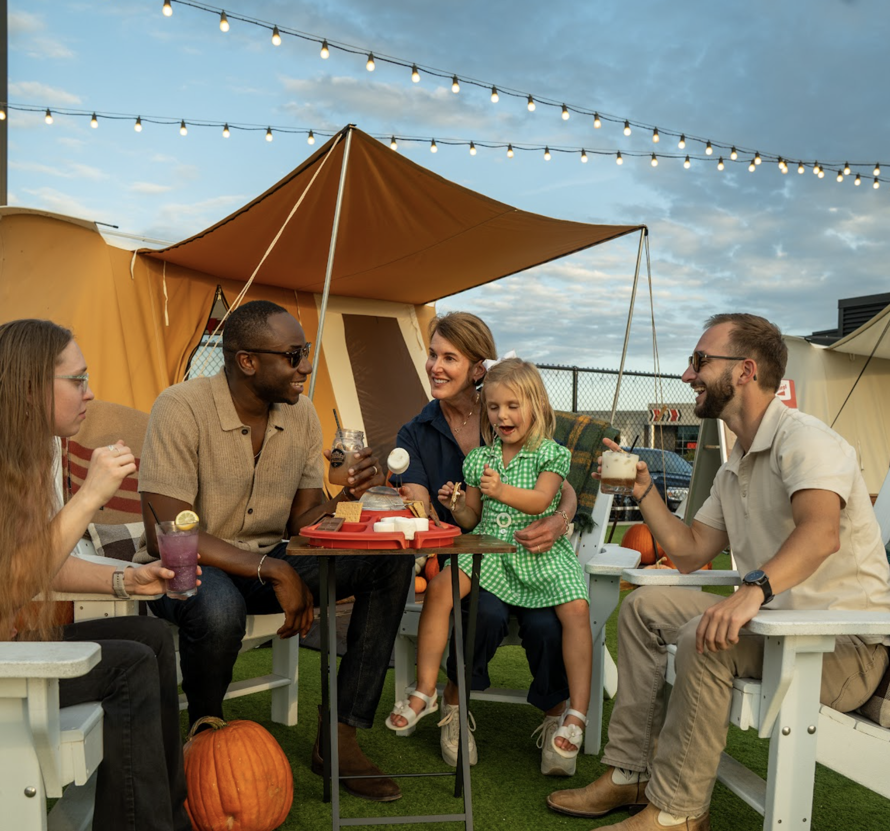 Five people, including a smiling child, sit outdoors around a table with drinks and snacks. They appear to be enjoying a casual gathering under string lights with tents and pumpkins nearby.