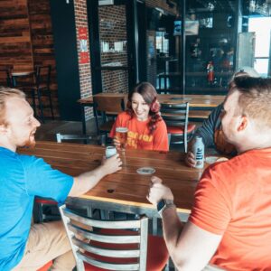 Three friends sit at a wooden table in a bar, laughing and holding drinks.