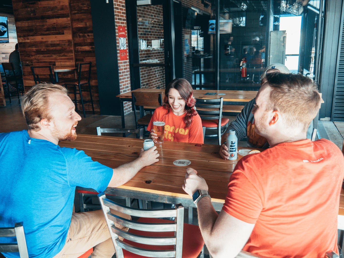 Three friends sit at a wooden table in a bar, laughing and holding drinks.