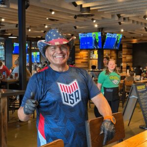 Smiling man wearing an American flag cowboy hat and USA shirt gives a thumbs-up inside Chicken N Pickle during the 10% Giveback Night Benefiting The Arya Foundation. TVs and diners are visible in the background.