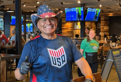 Smiling man wearing an American flag cowboy hat and USA shirt gives a thumbs-up inside Chicken N Pickle during the 10% Giveback Night Benefiting The Arya Foundation. TVs and diners are visible in the background.
