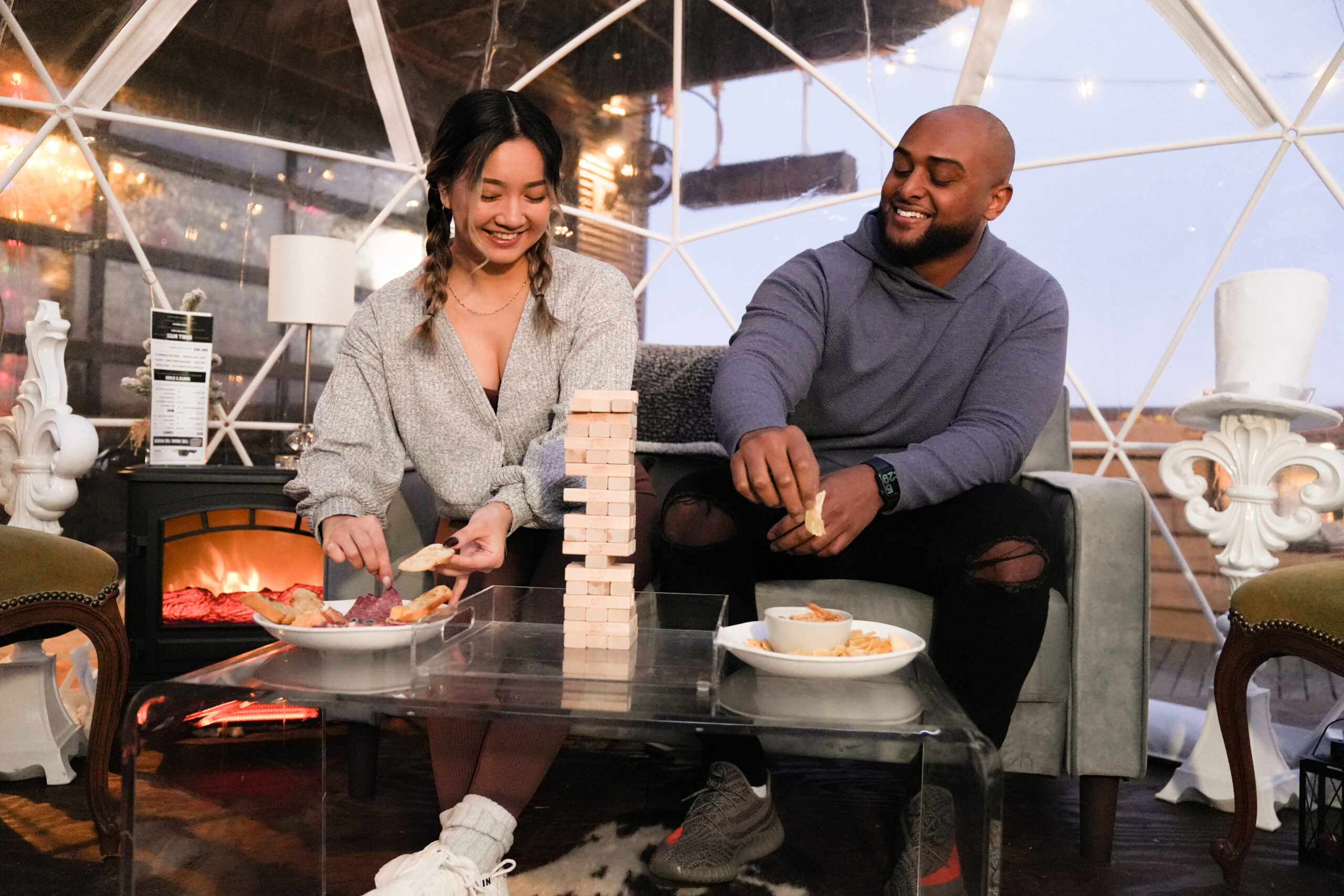 A man and woman play a block-stacking game together inside a lit outdoor dome while eating snacks.