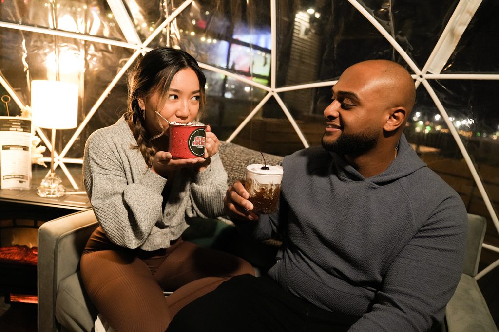 A man and woman sitting inside a transparent outdoor dining dome, both holding cocktails.