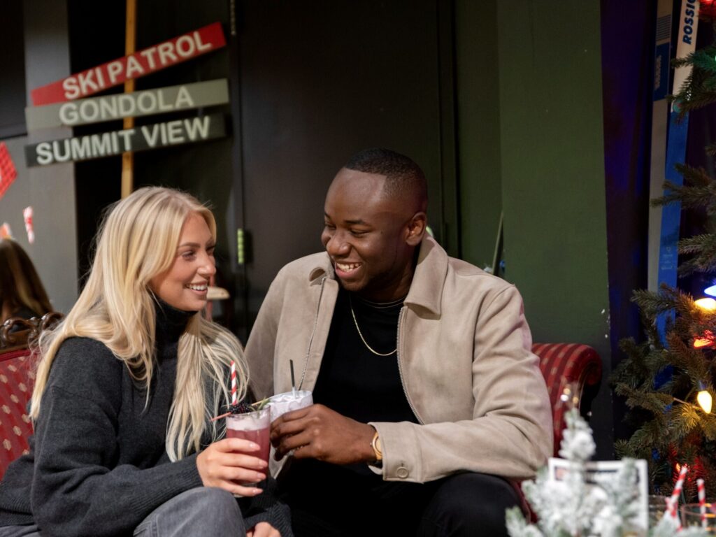 A man and a woman sitting on a couch smile at each other while holding cocktails in a decorated holiday setting.