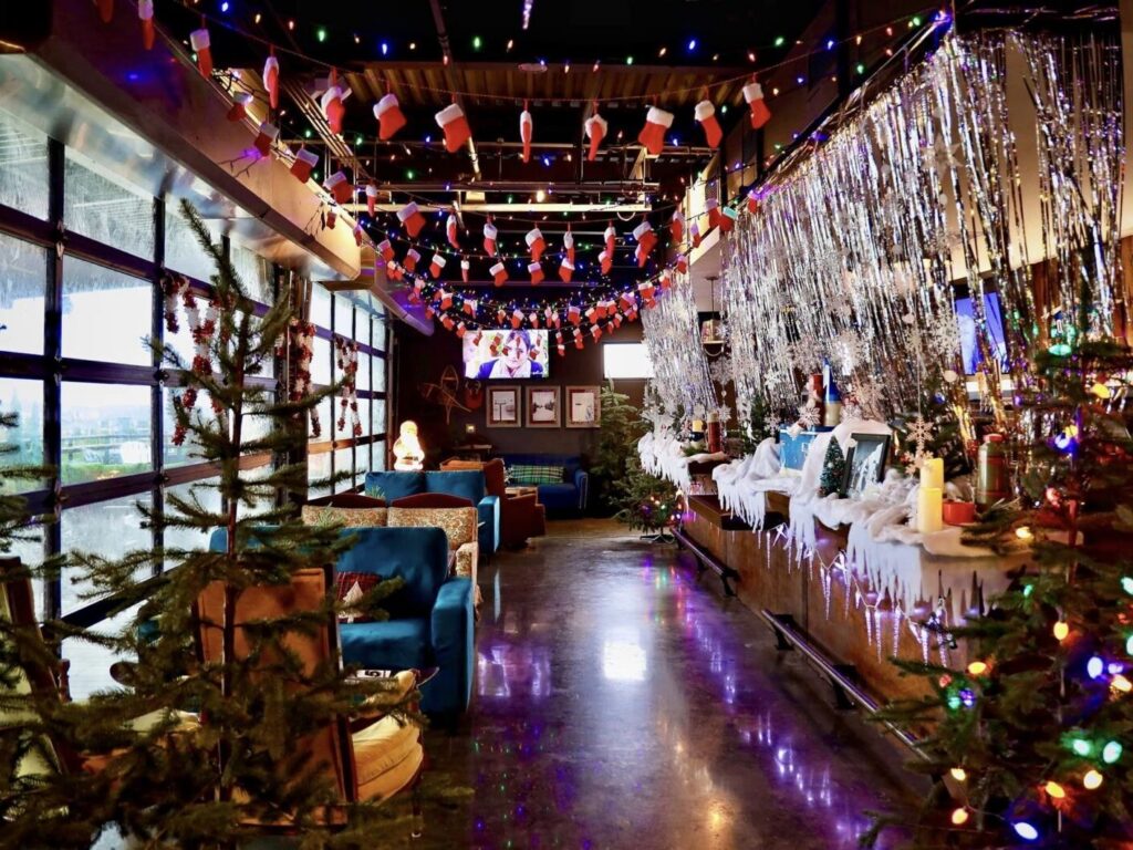 A restaurant interior decorated for Christmas with strings of stockings, colorful lights, silver fringe curtains, and several small pine trees.