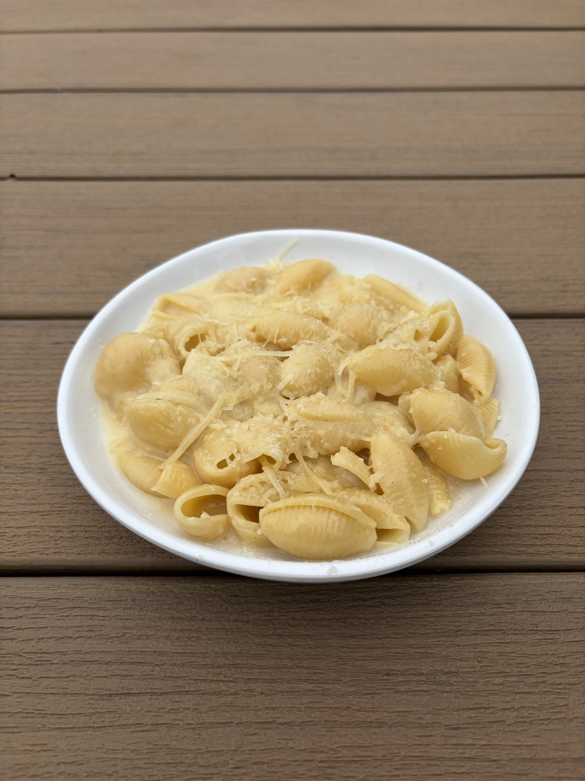 A white bowl filled with shell pasta covered in a creamy sauce and topped with grated cheese sits on a wooden table.